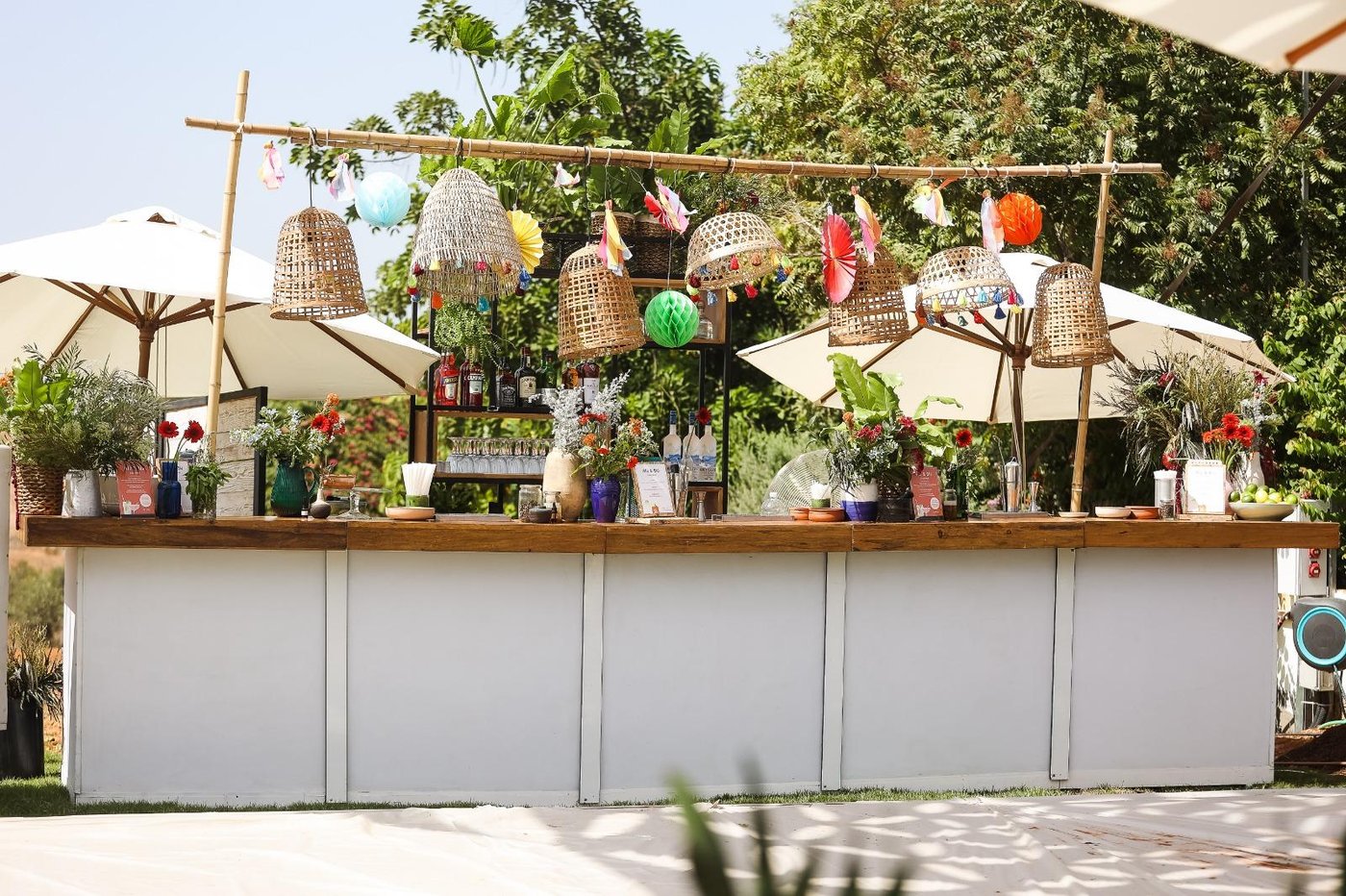 An outdoor boho-style event bar setup with woven pendant lampshades, colorful paper fans and lanterns, string decorations, umbrellas, and greenery