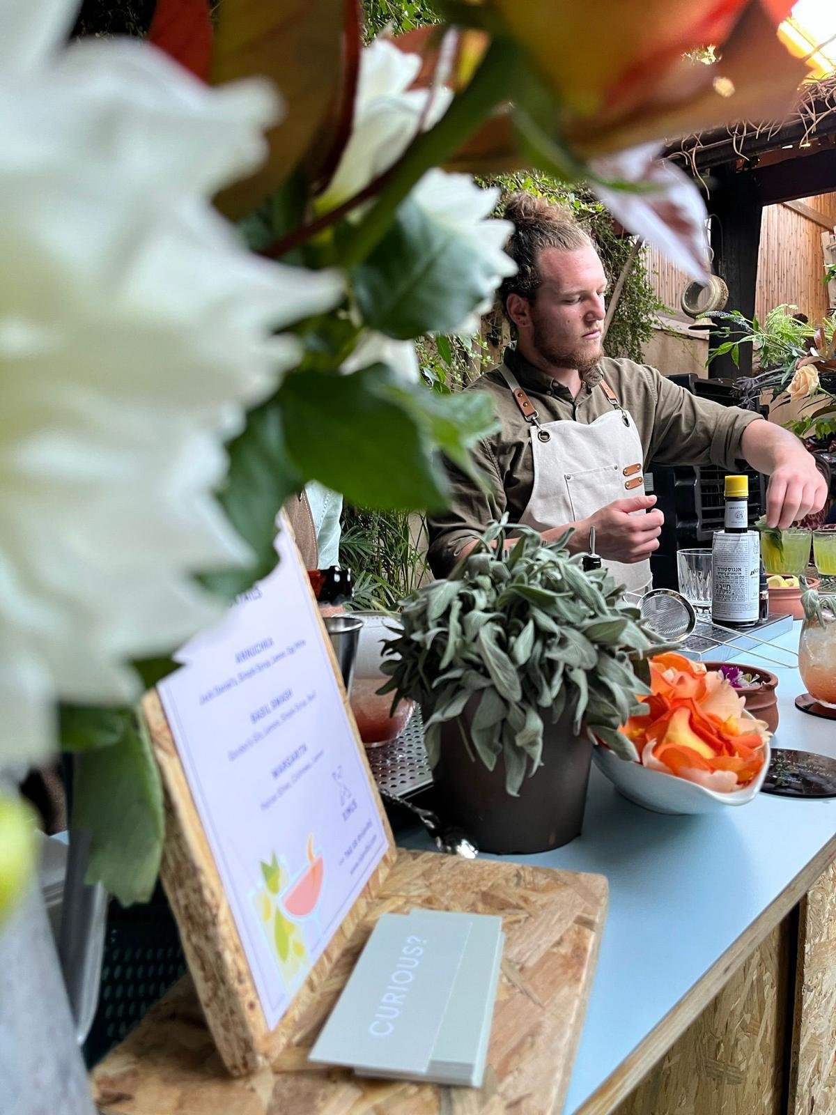 A bartender preparing cocktails at an outdoor event bar, surrounded by flowers and a printed cocktail menu